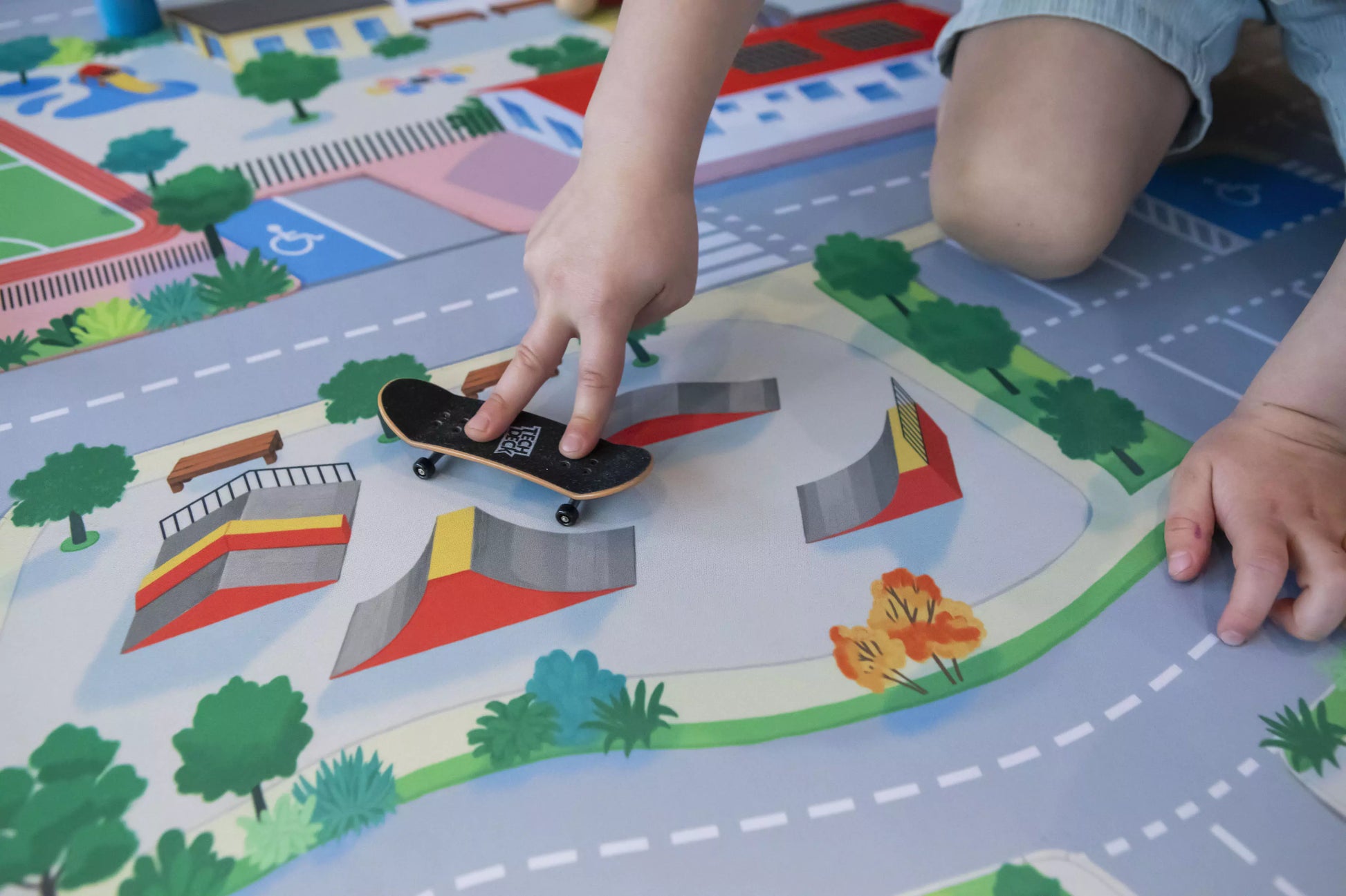 A child dives into imaginative play with a fingerboard on "The Community" Town Play Mat by Habitat Mat, which features a lively skatepark design. Using their fingers, the child performs skateboarding tricks on the vibrantly illustrated ramps and rails.