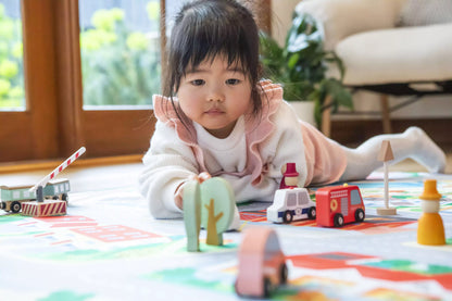 A young child with long dark hair lies on "The Community" Town Play Mat by Habitat Mat, deeply absorbed in imaginative play with wooden toy vehicles and buildings. Natural light streams through a nearby window, casting a gentle glow, and vibrant green plants can be seen in the background.