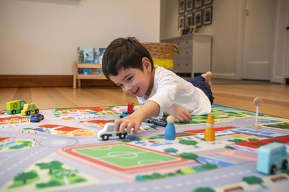 A young boy, radiating happiness, lies on the floor in a cozy, well-lit room as he embarks on imaginative play with toy cars across "The Community" Town Play Mat by Habitat Mat, depicting a vibrant city scene complete with roads and buildings, crafting his own miniature community.