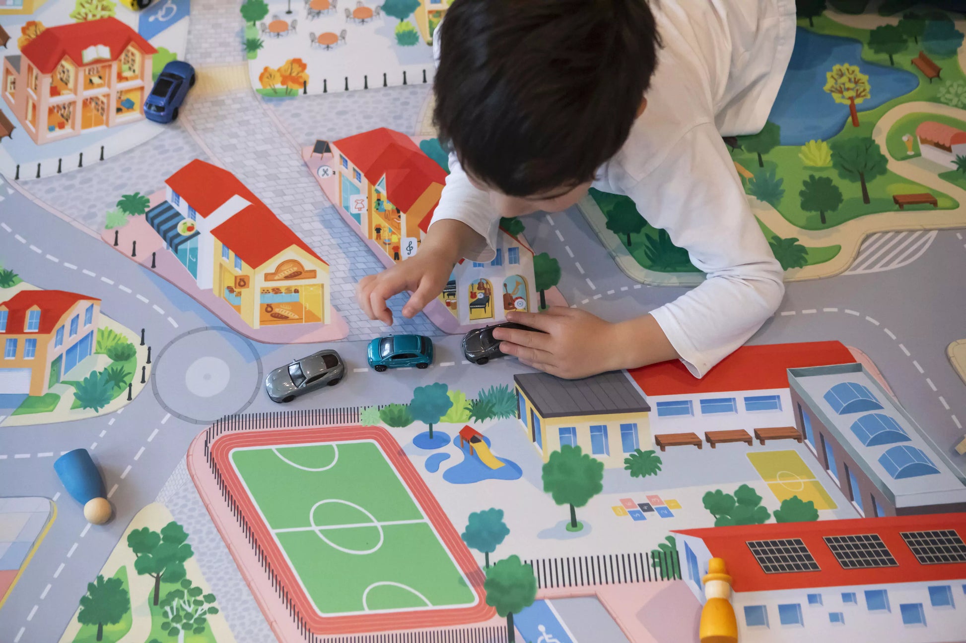 A child with short dark hair and a white shirt immerses in imaginative play, driving toy cars across Habitat Mat's "The Community" Town Play Mat, which unfurls into a vibrant miniature town featuring roads, buildings, a soccer field, and a park.