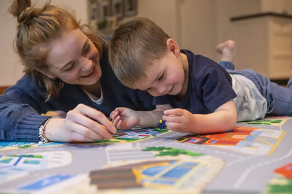 A woman and child are lying on the floor, engaging in imaginative play with toy cars and figures on "The Community" Town Play Mat by Habitat Mat. Both are smiling, clearly absorbed in the fantasy world they've created. The room appears cozy and well-lit.