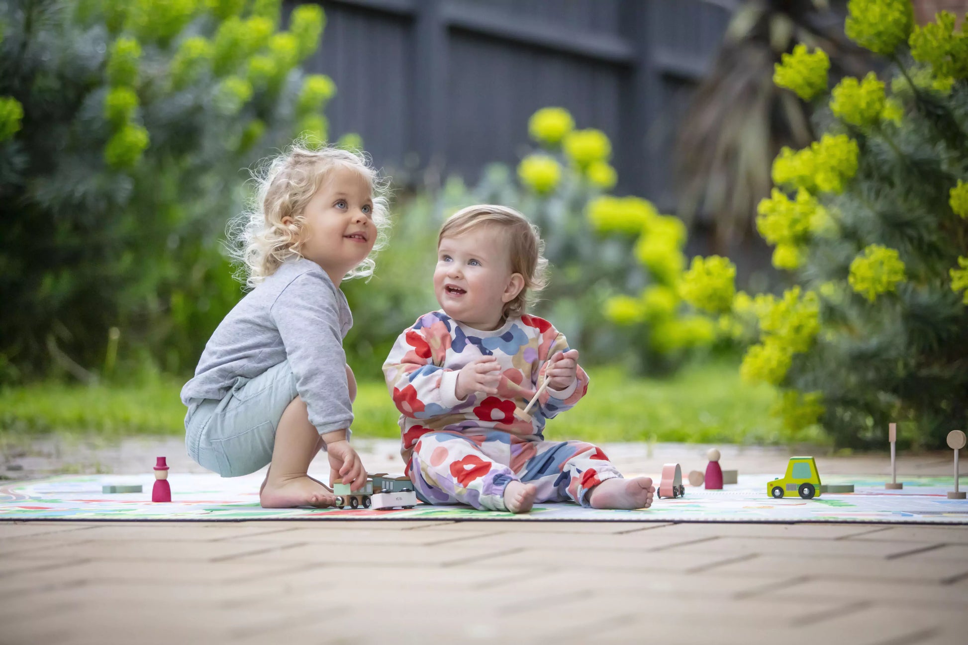 Two young children are enjoying themselves on "The Community" Town Play Mat by Habitat Mat outdoors. One child with curly hair is crouching in a gray shirt and blue shorts, while the other child sits nearby in a vibrant outfit. Engaged in imaginative play, they are surrounded by lush greenery and toys, with yellow flowers accentuating the background.