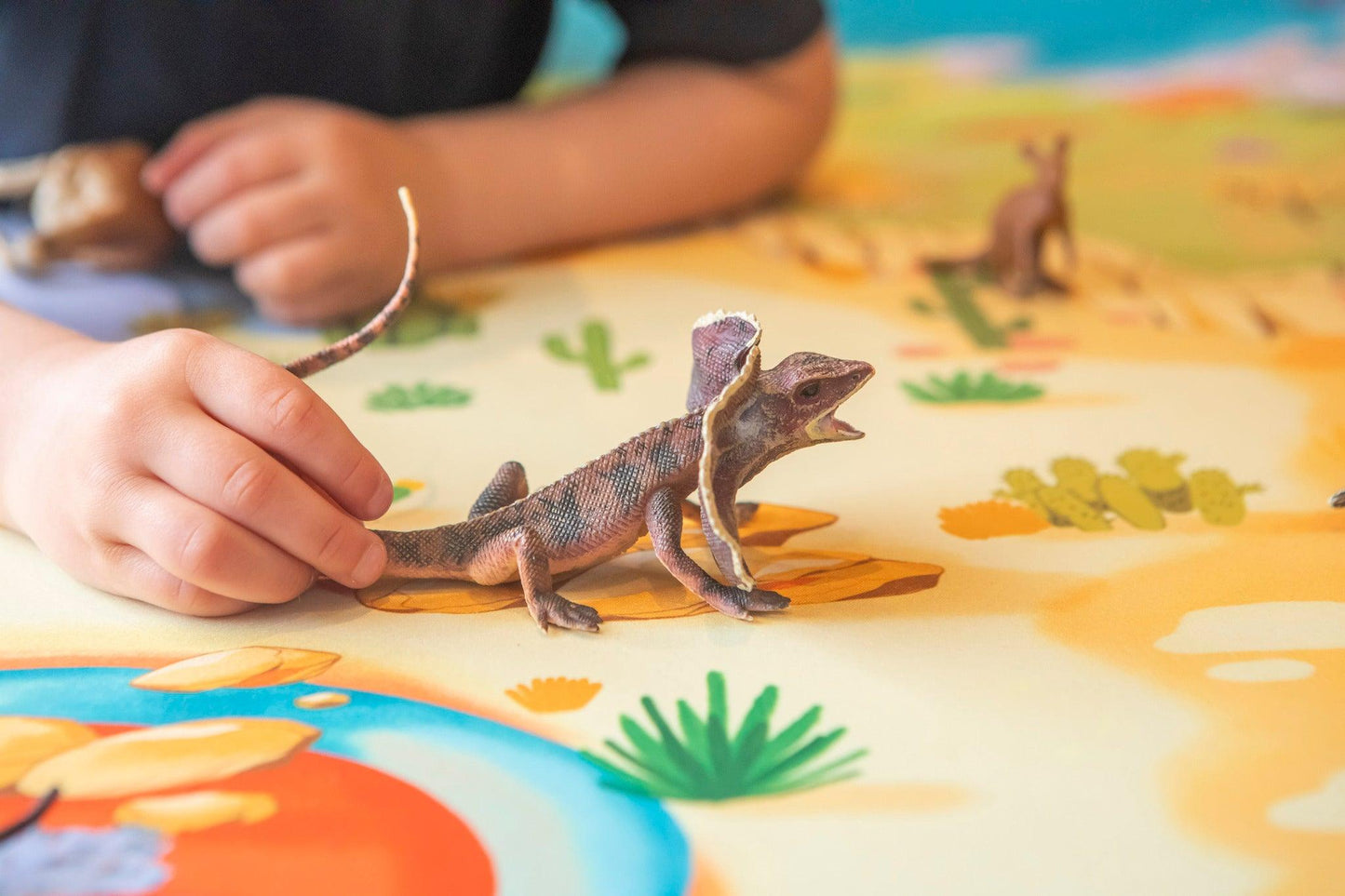 A child plays with a toy lizard on "The Explorer" Wildlife Play Mat from Habitat Mat, which features vibrant desert-themed illustrations with cacti and rocks. The toy lizard has an open mouth and extended frill. Nearby, the child's hands engage with other small animal toys on the wildlife play mat.