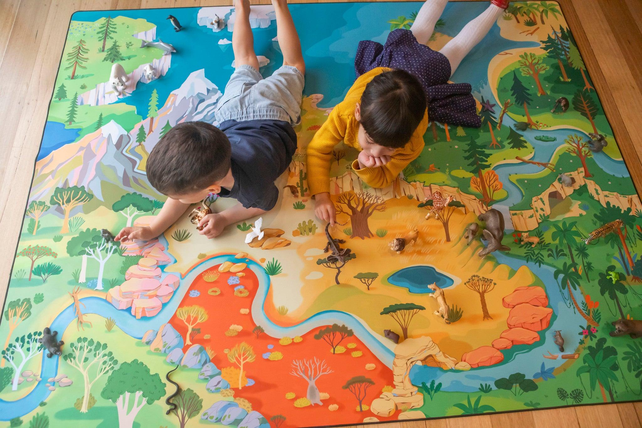 Two children, one in a yellow shirt and the other in a blue one, are lying on their stomachs, playing on "The Explorer" wildlife play mat by Habitat Mat. Surrounded by toy animals, they engage in imaginative play across various landscapes and animals, embarking on new adventures as little explorers.