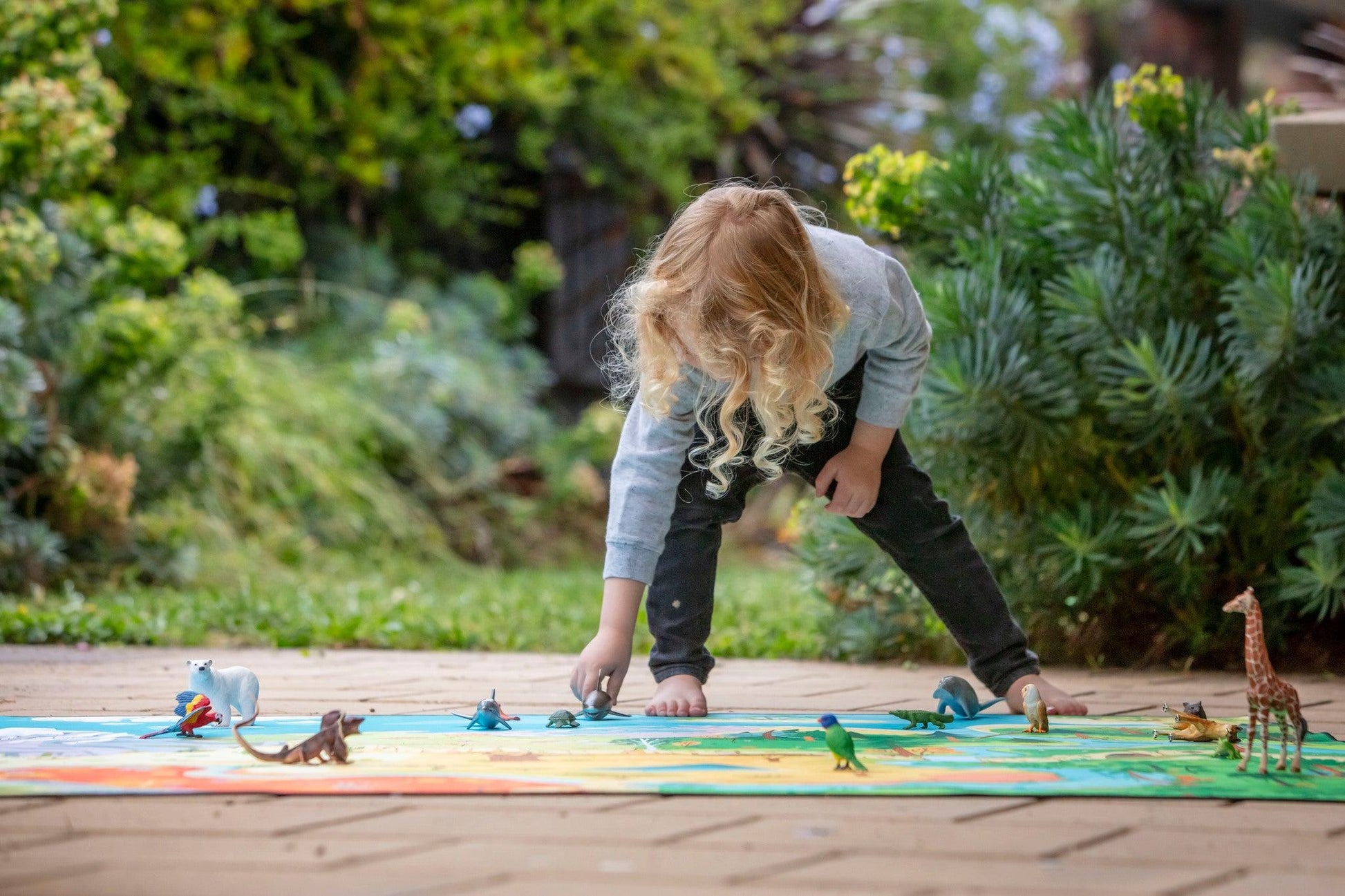 On a vibrant "The Explorer" Wildlife Play Mat by Habitat Mat, a young child with long curly hair enjoys playing with toy dinosaurs outdoors. Lush greenery envelops the patio area, forming a natural backdrop.