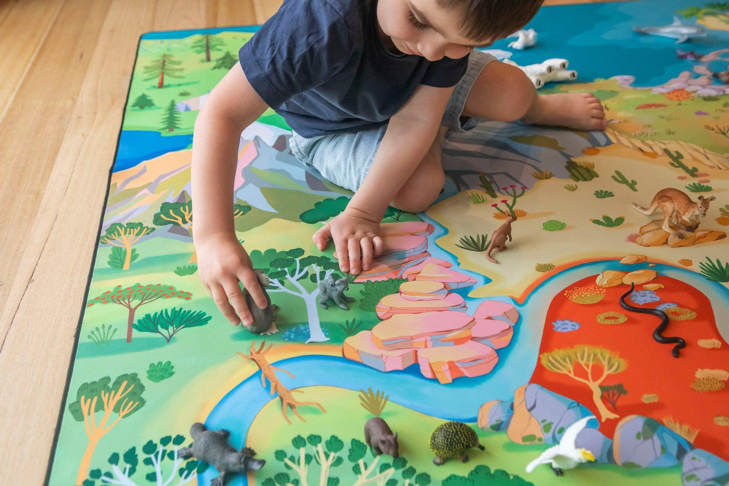 Dressed in a blue shirt and shorts, the child dives into a world of adventure on Habitat Mat's "The Explorer" Wildlife Play Mat, strategically placing animal figurines across vibrant forest landscapes and rocky terrains, unified by a meandering river.