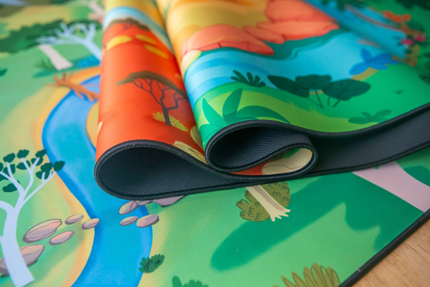 The Explorer" Wildlife Play Mat by Habitat Mat showcases a vibrant landscape featuring green trees, a blue river, rocks, and orange hills. Partially rolled to reveal its layers, the colorful illustrated play mat is set on a light wooden surface.