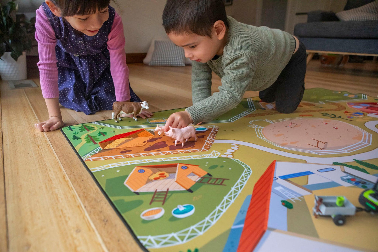 Two young children are engaged in imaginative play on a vibrant "The Farmer" Farmyard Play Mat by Habitat Mat. They use toy farm animals, such as a cow and pig, as they explore the neoprene mat's colorful farm scenes featuring houses and fields. The room's wooden flooring and a couch in the background create the perfect backdrop for their adventures.