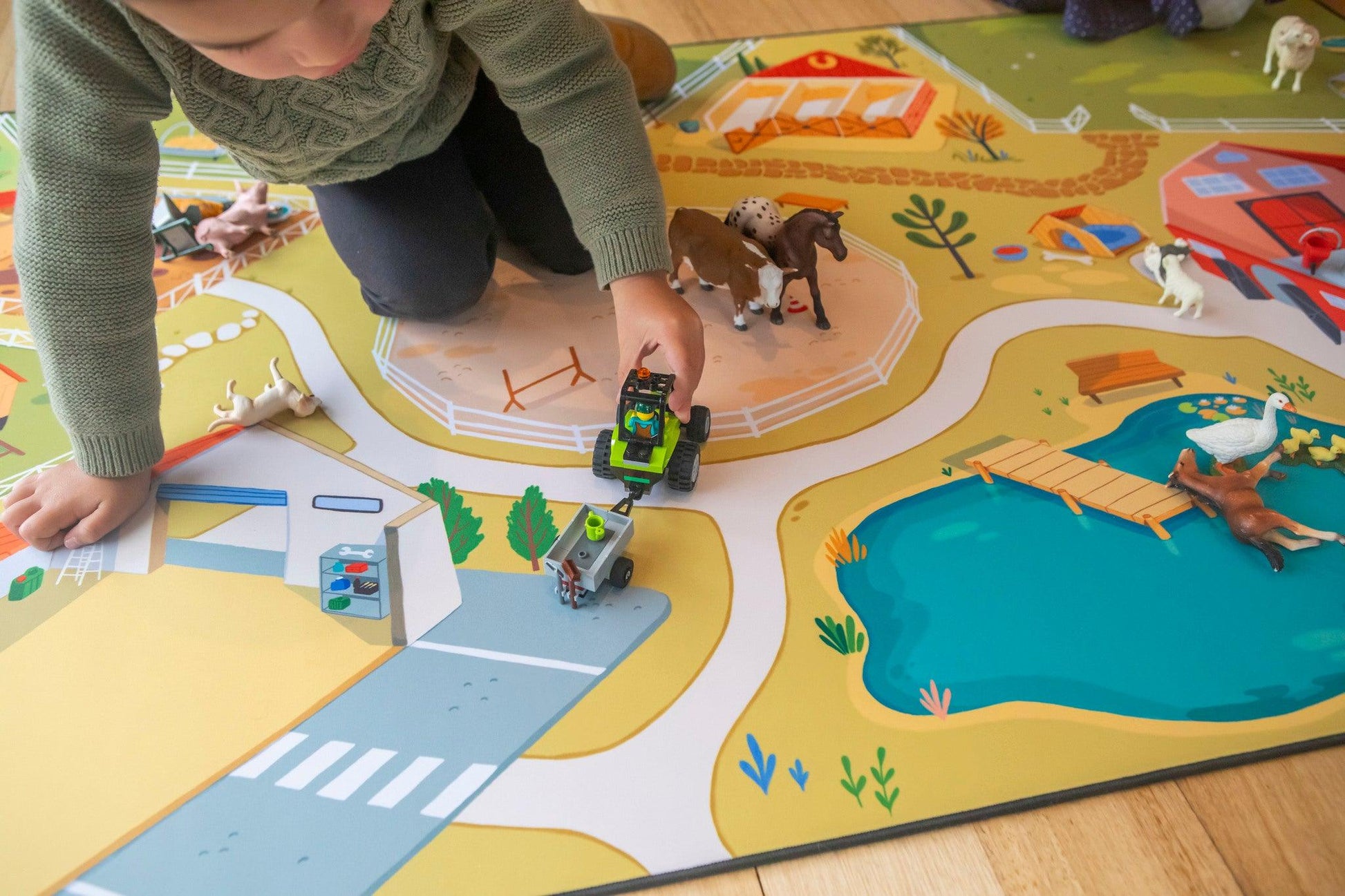 Kneeling on "The Farmer" farmyard play mat, a child in a green sweater is deeply engaged in the vibrant farm scene. They maneuver a toy tractor between small animals, navigating roads, a pond, and charming farm buildings depicted on the colorful Habitat Mat.