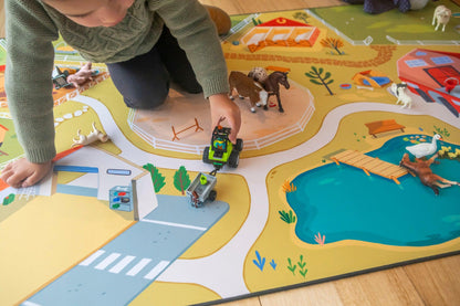 Kneeling on "The Farmer" farmyard play mat, a child in a green sweater is deeply engaged in the vibrant farm scene. They maneuver a toy tractor between small animals, navigating roads, a pond, and charming farm buildings depicted on the colorful Habitat Mat.
