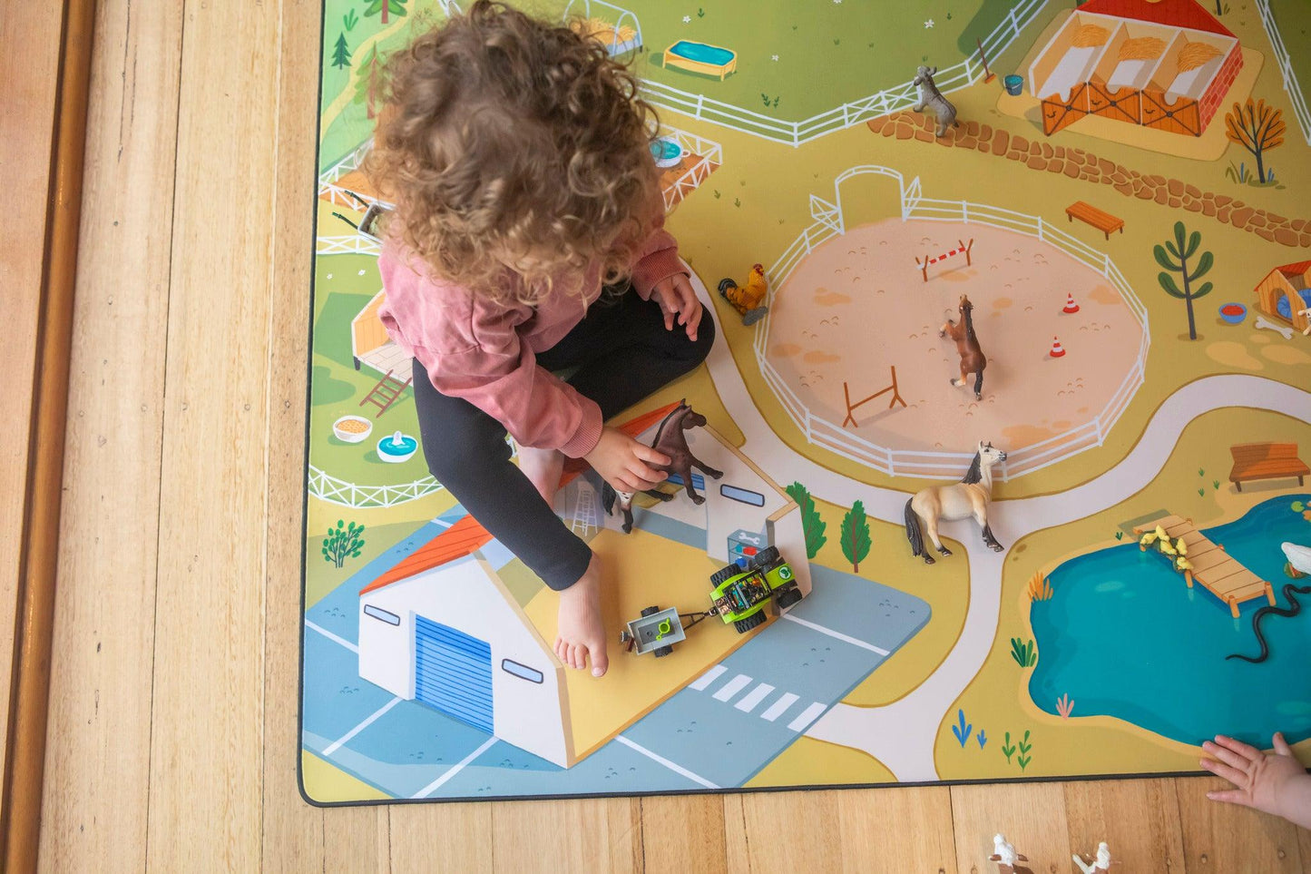 A child with curly hair is sitting on a vibrant "The Farmer" Farmyard Play Mat from Habitat Mat, playing with toy animals and a tractor. The neoprene mat showcases illustrations of a farm complete with a pond and buildings, as the child focuses intently on arranging the toys.