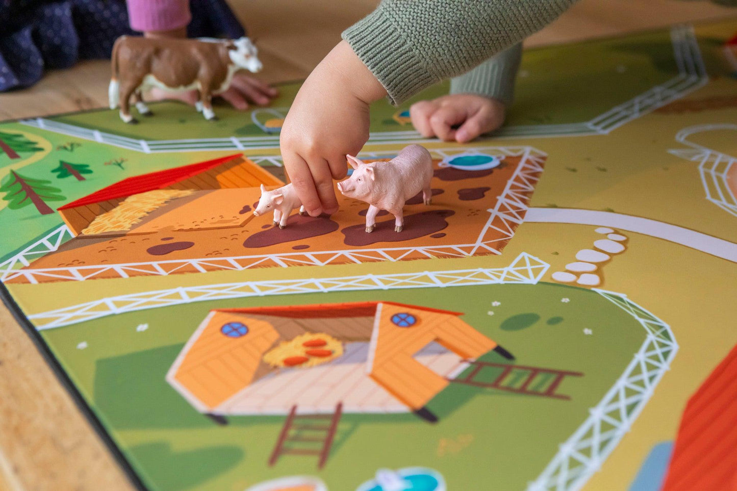 A child plays on "The Farmer" Farmyard Play Mat by Habitat Mat, featuring bright farm elements such as a barn and path. Their small hand interacts with a pig figurine in this neoprene farmyard setting, sparking endless imagination.