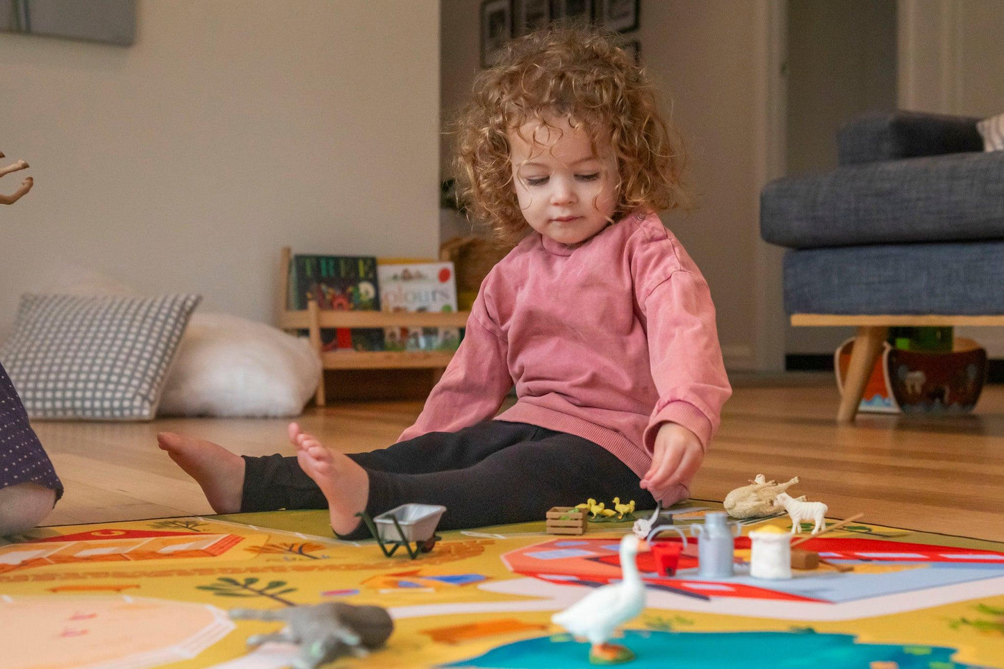 A young child with curly hair sits on a wooden floor, dressed in a pink sweater and black pants. They are joyfully playing on "The Farmer" farmyard play mat by Habitat Mat, surrounded by animal toys. In the background, a gray couch and children's books can be seen on a low shelf.