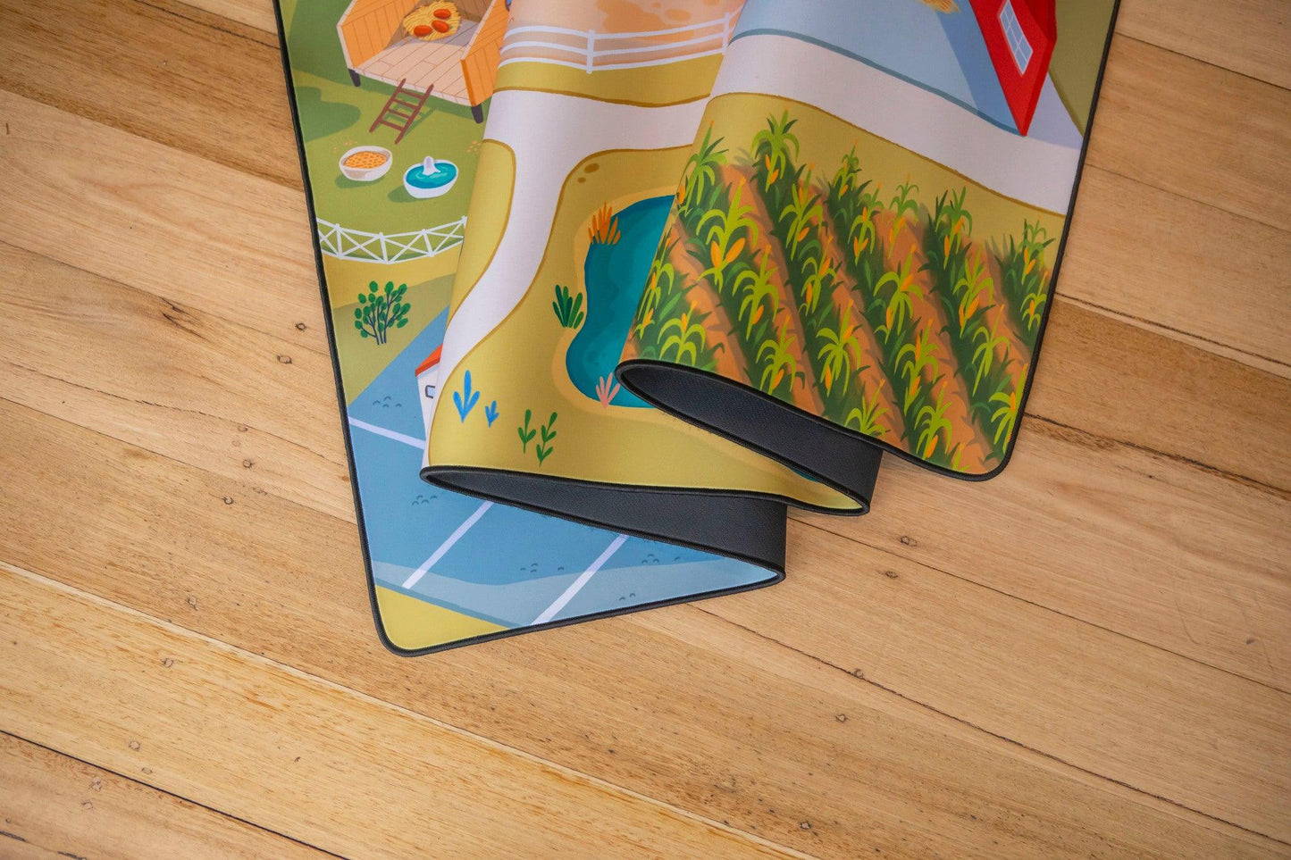 A vibrant "The Farmer" Farmyard Play Mat from Habitat Mat adorns the wooden floor, displaying vivid farm-themed illustrations. Crafted from durable neoprene, this mat features charming elements like a barn, fields, and a pond. It's partially rolled up, enticing you to explore more of its delightful design.